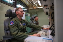 Two men in green military flight suit uniforms sit at a console table inside a launch control center capsule.