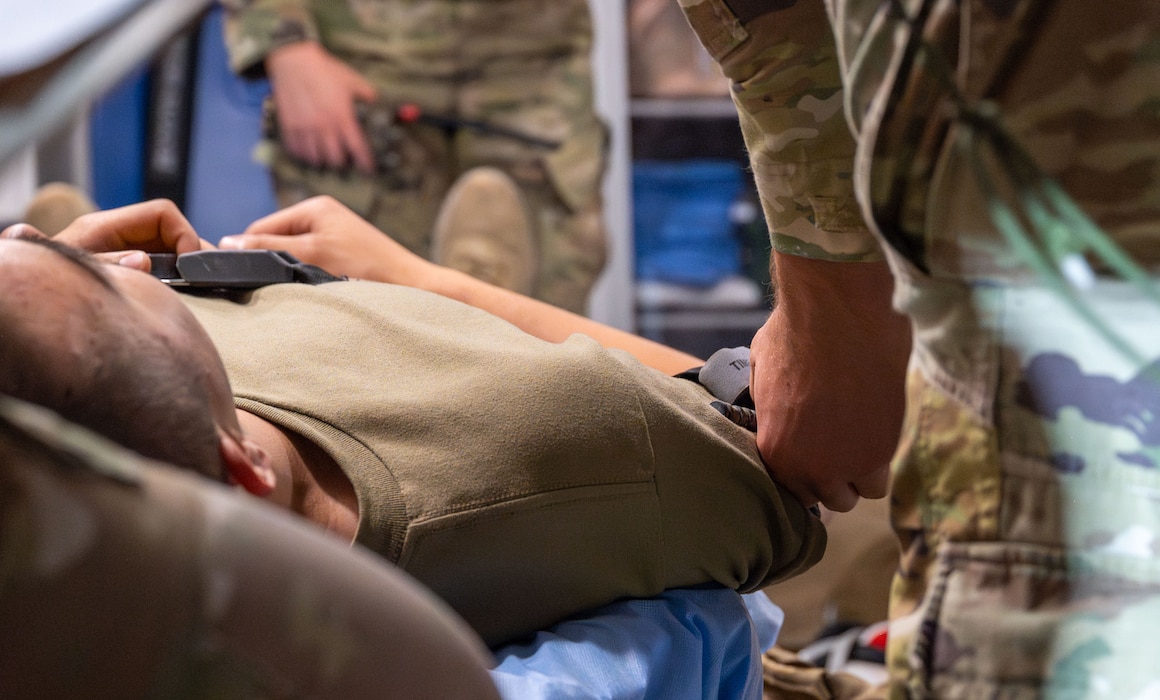 A man tightens a tourniquet on another man's arm who is laying on a gurney in an ambulance.