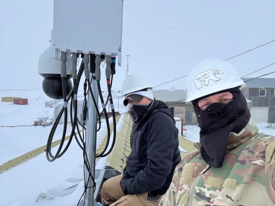Two men in cold-weather gear pose near communications equipment.