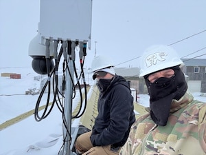 Two men in cold-weather gear pose near communications equipment.