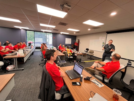 a group of individuals in red shirts are seated in rows at tables with one individual in a dark shirt stands at the front of the room talking to them.