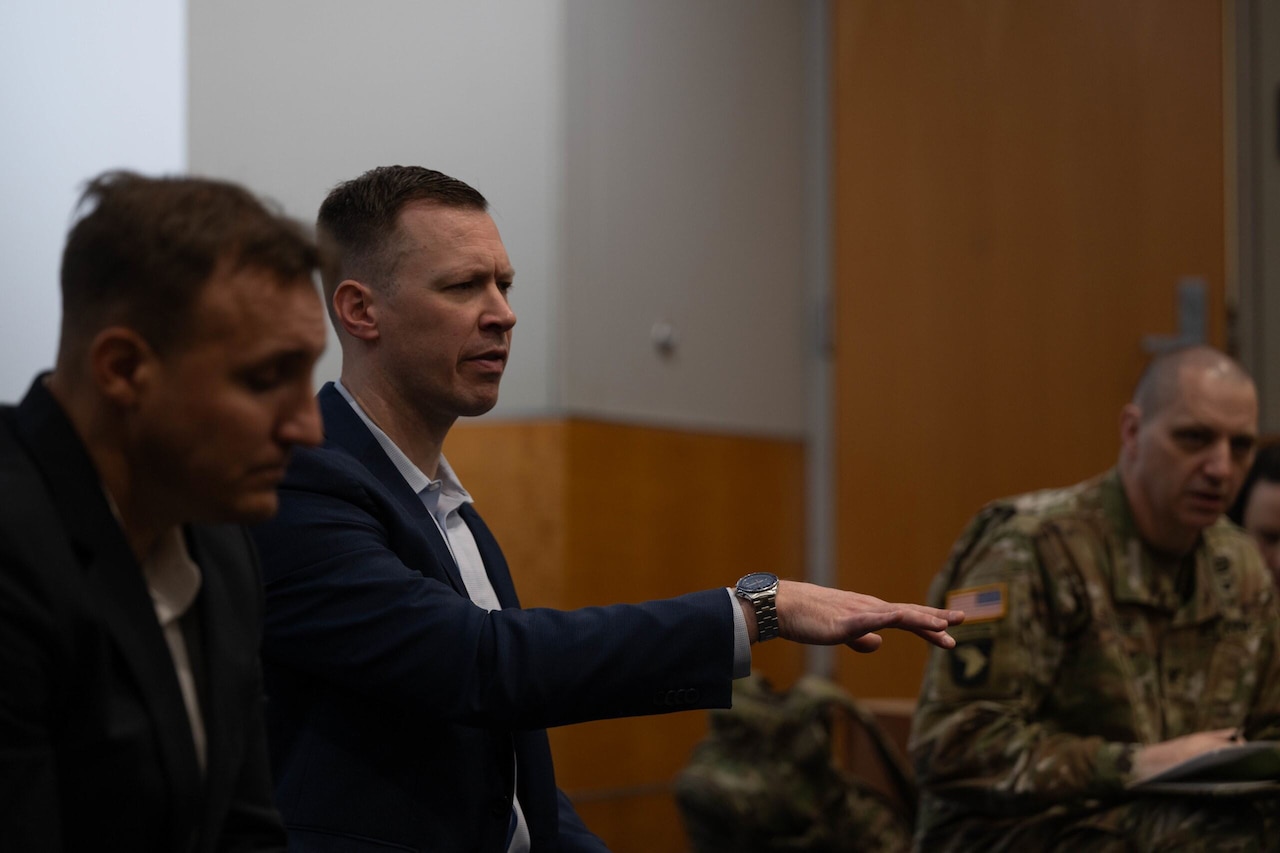 A man in a business suit is in a room, gesturing with his right hand. A man in a camouflage military uniform is in the background looking at him.