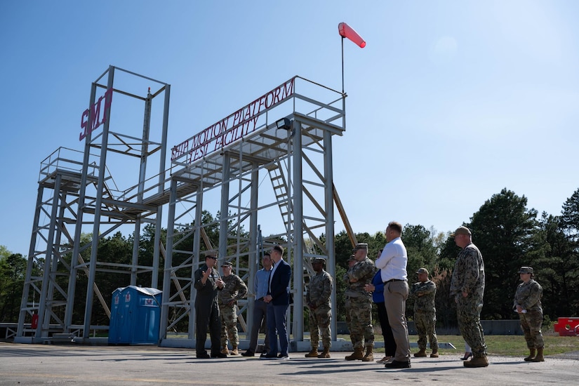Roughly one dozen people, most in camouflage military uniforms and a few in civilian business attire, are standing outdoors, gathered around a large, scaffolding-like metal structure.