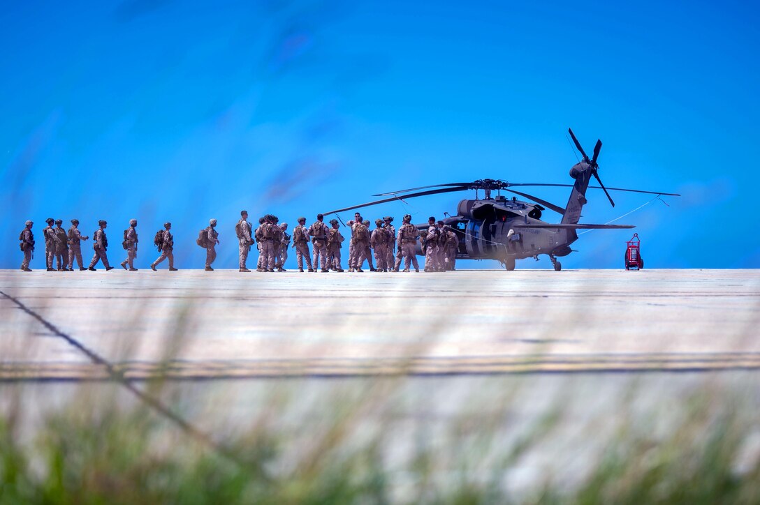 Marines and soldiers  walk along a beach toward a helicopter with a blue sky in the background.