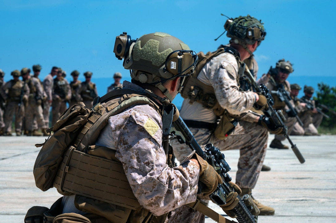 Marines holding rifles kneel on the ground in a row as others stand and look on in the background.