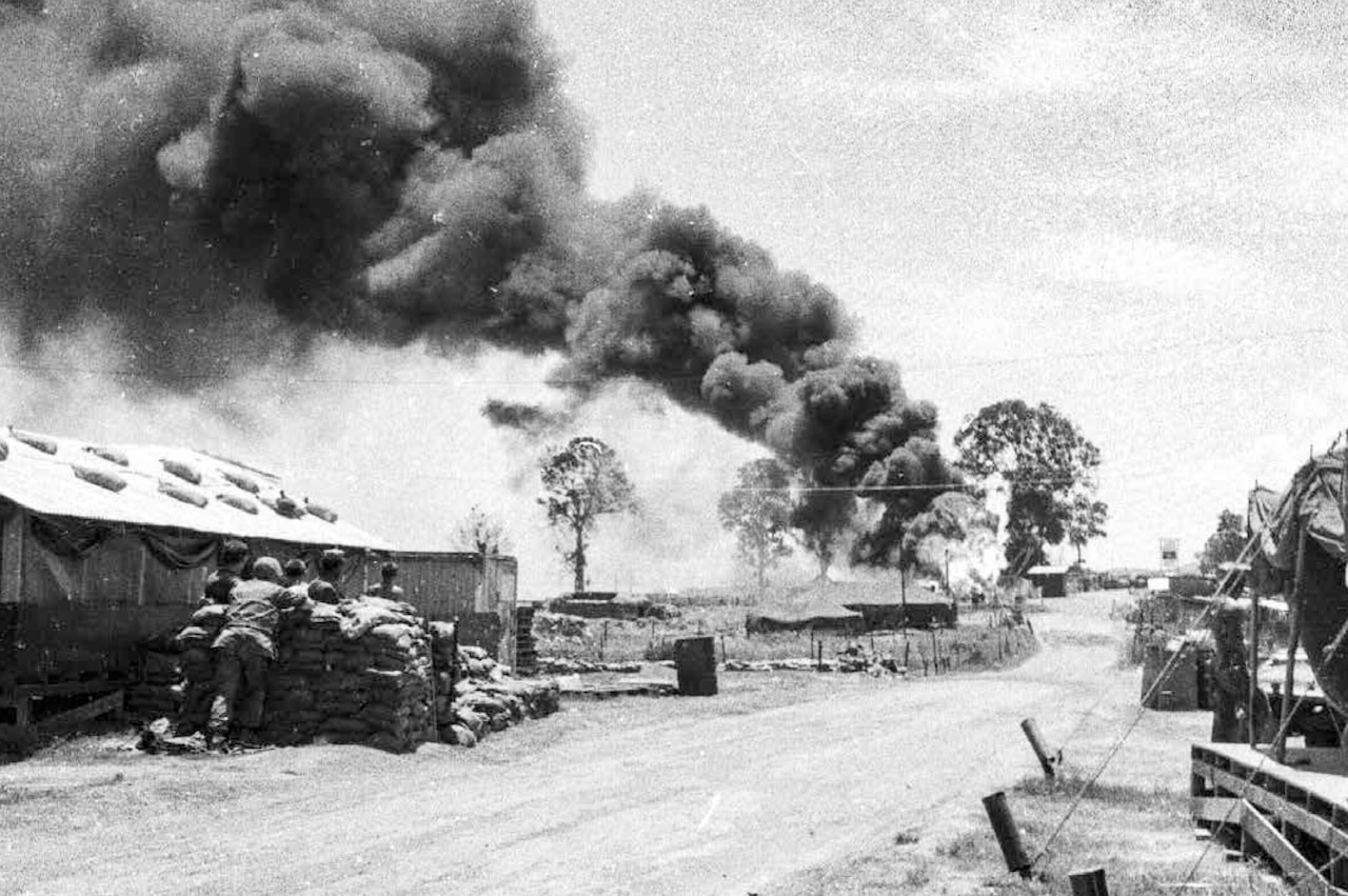 Men behind sandbags near a building and tents watch a black plume of smoke rise from the ground in the distance.