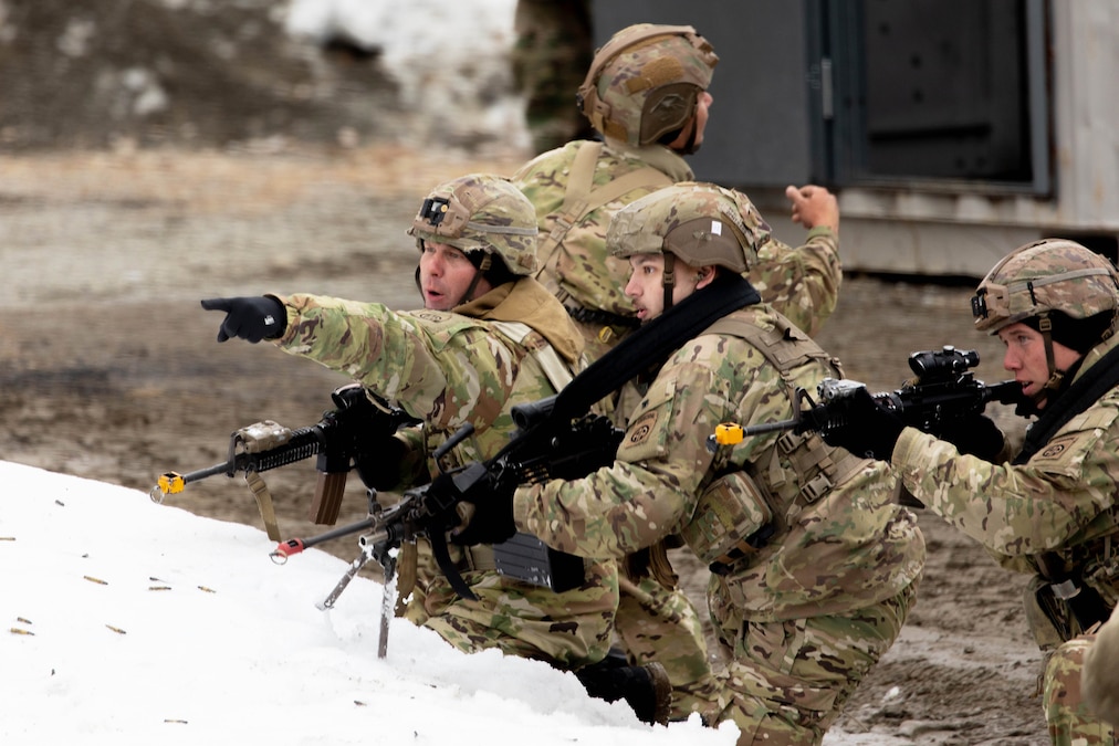 Troops in tactical gear kneel in the dirt while aiming weapons over a pile of snow covered in ammo.