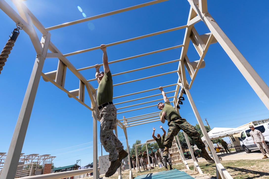 Marines climb across monkey bars as fellow Marines watch against a blue sky.