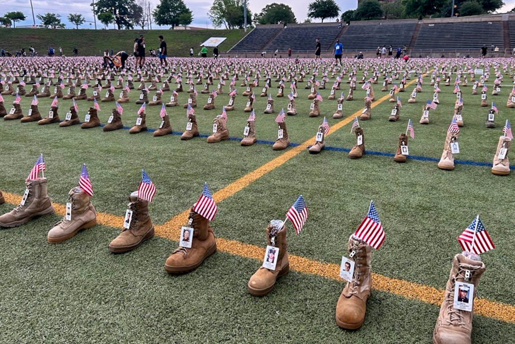 Thousands of boots with an ID and an American flag sit on a turf field as people walk around.
