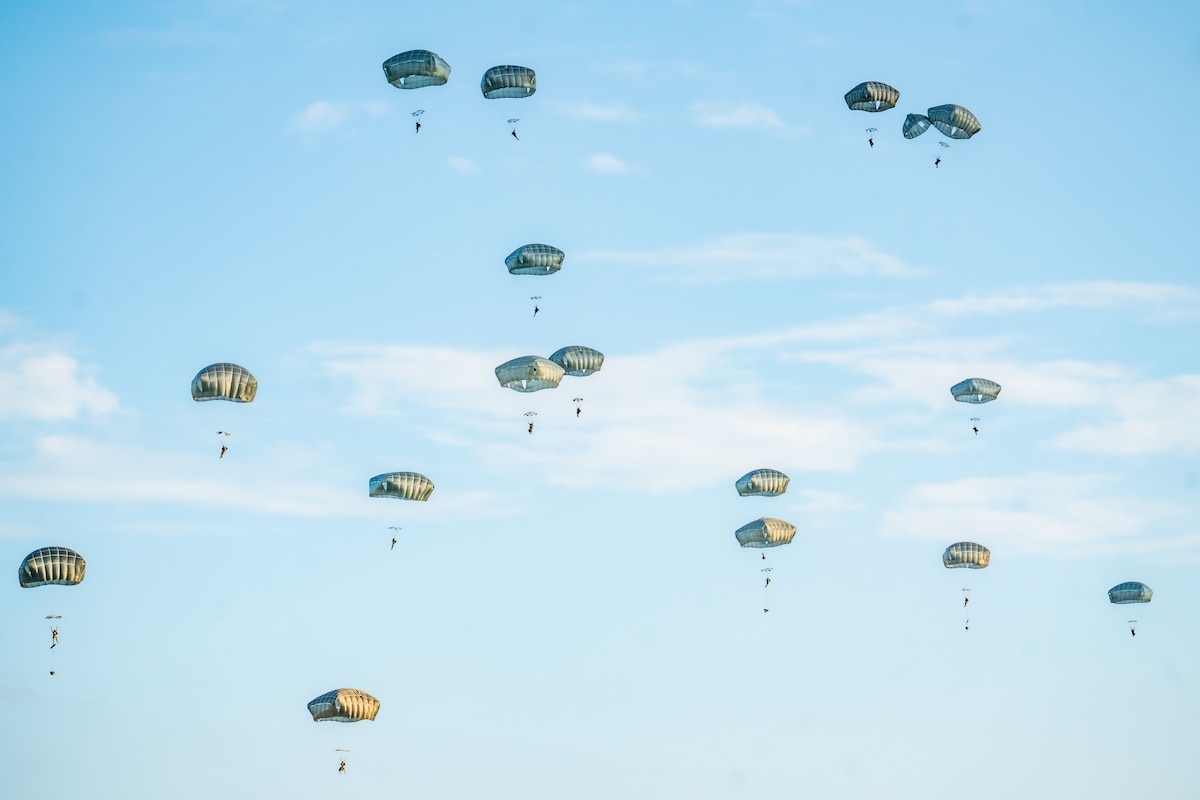 More than a dozen paratroopers descend by parachute in a blue sky.