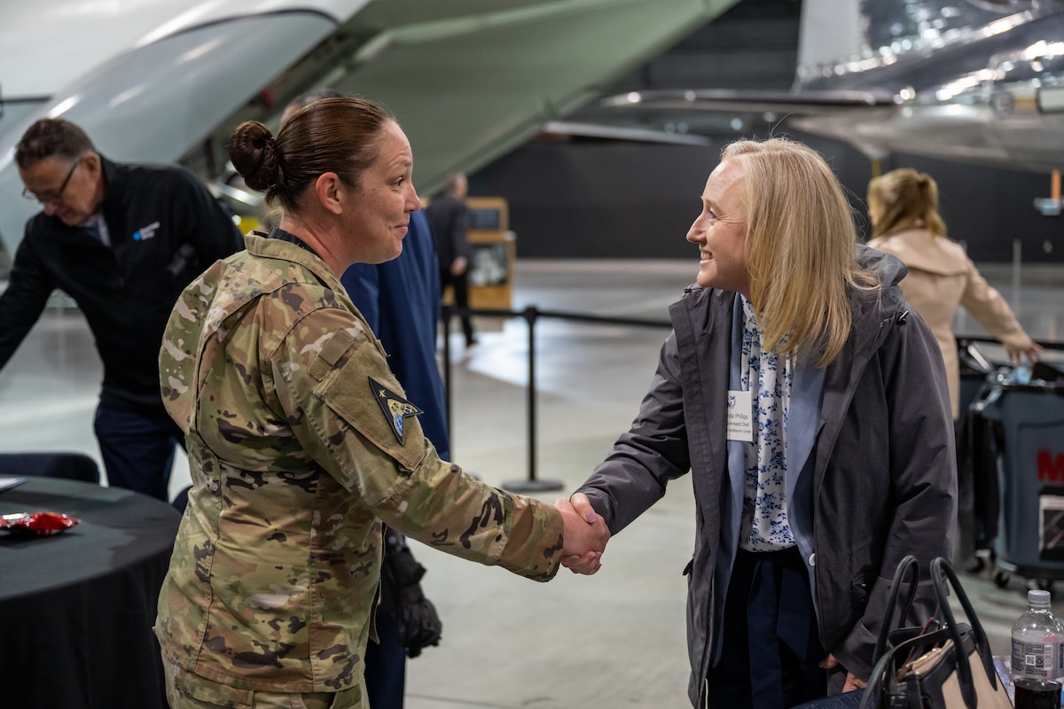 A woman, in camouflage military uniform, shakes the hand of a woman, in civilian business casual attire,