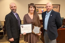 Three people pose for a photo, the person standing in the middle is holding two awards.
