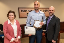 Three people pose for a photo, the person standing in the middle is holding two awards.