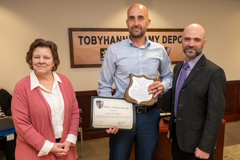 Three people pose for a photo, the person standing in the middle is holding two awards.
