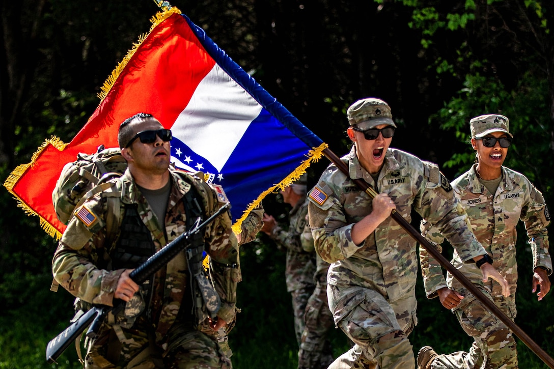 A soldier runs with a rifle as two others, one carrying a flag, run to the right and cheer.