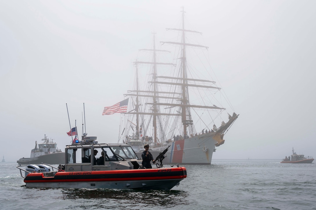 A Coast Guard cutter and three smaller vessels move through a body of water on a foggy day.