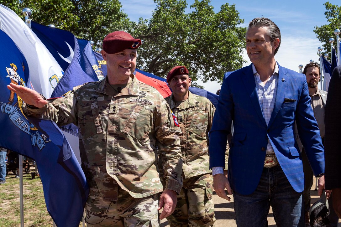 A man in a suit walks and talks next two two soldiers outside on a sunny day.