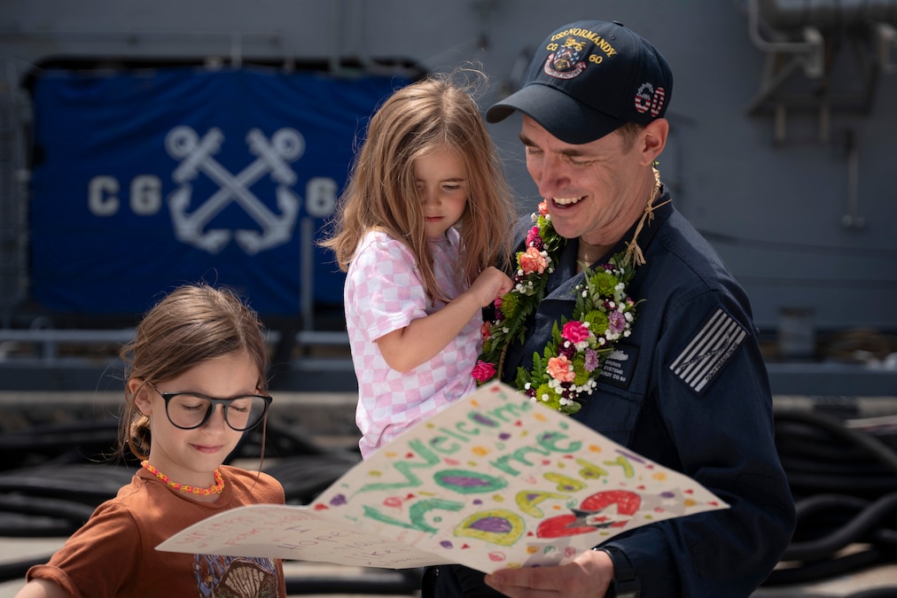 A smiling sailor holds one child and stands next to another as all three look at a homemade card.