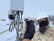 Two men in cold-weather gear pose near communications equipment.