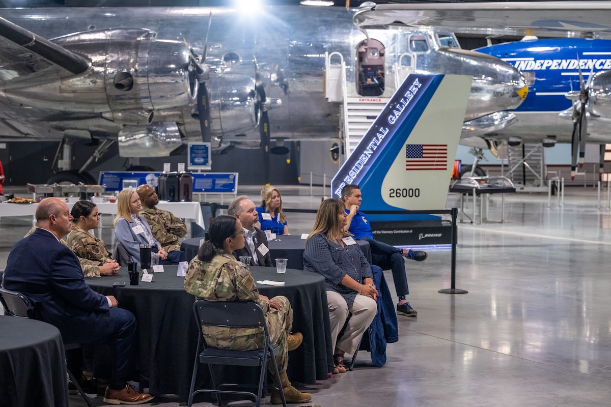 A large group of people, both military in uniform and civilians in business casual, sit at round tables with black tablecloths, in front of a large sign saying Presidential Gallery and large silver aircraft