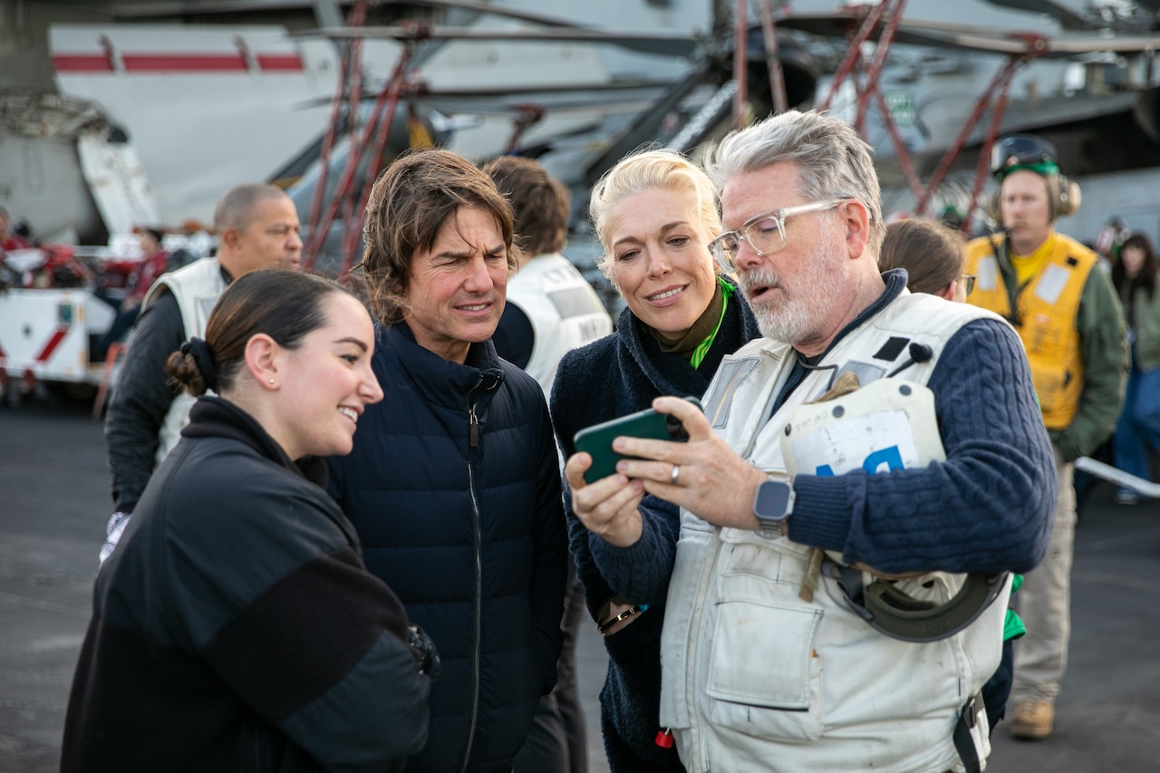 Four people look at a smartphone together while others mill around a flight deck behind them.