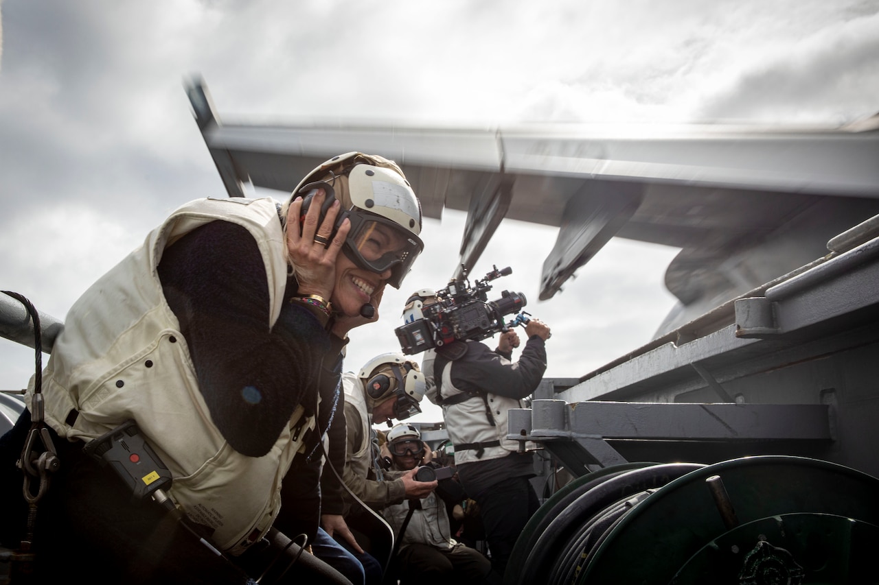 A woman in goggles and a helmet covers her ears as an aircraft takes off past her on a flight deck. A man in the background films it with a production camera.