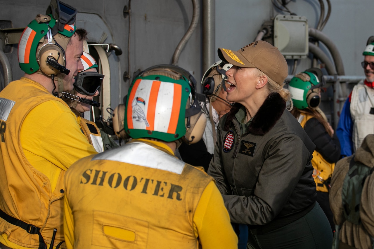 A woman talks with three men in bright yellow jackets and helmets. Others mill around behind them.