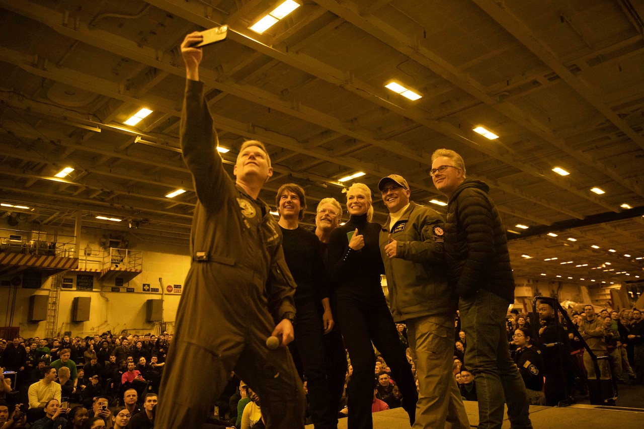 Six people standing on a stage take a selfie. Hundreds of people offstage watch them.