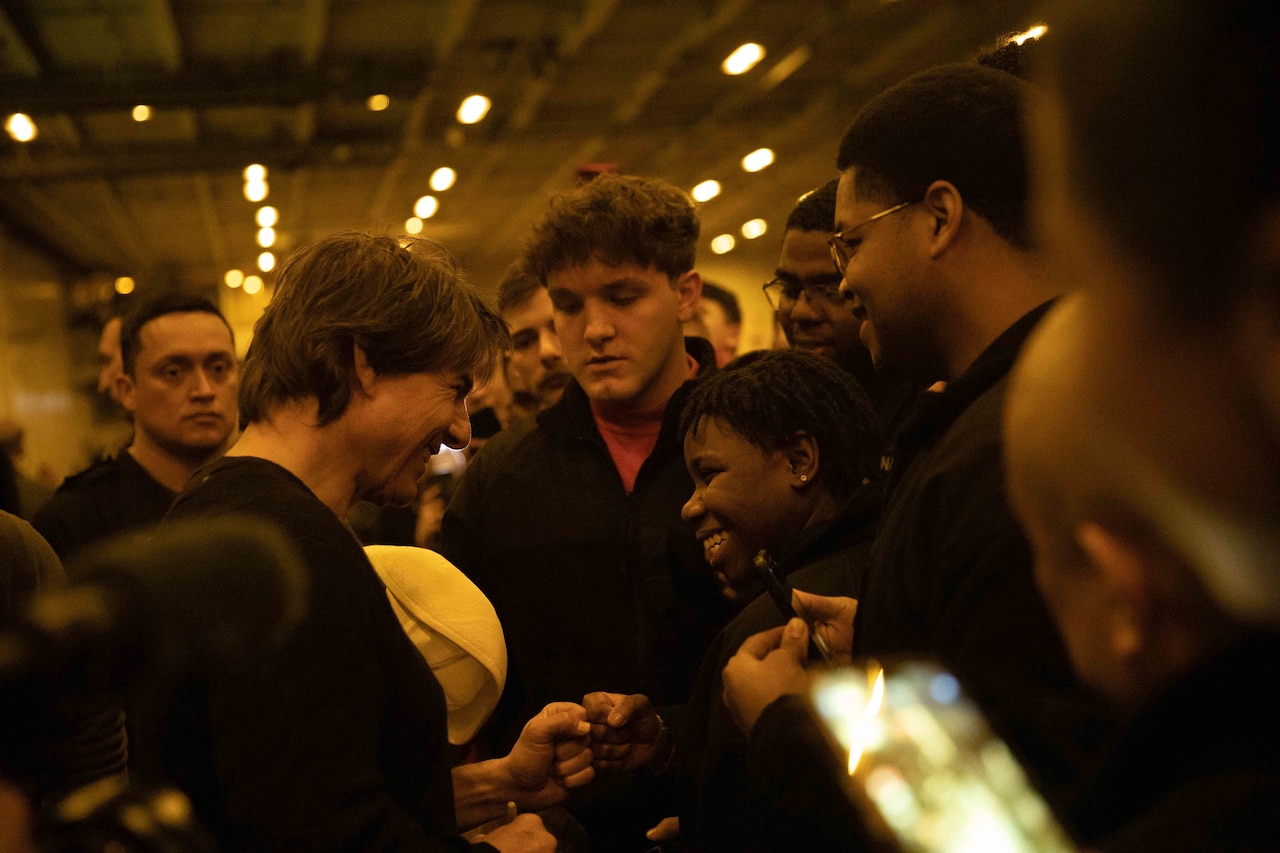 Several people crowd around a man, who fist bumps a smiling sailor.