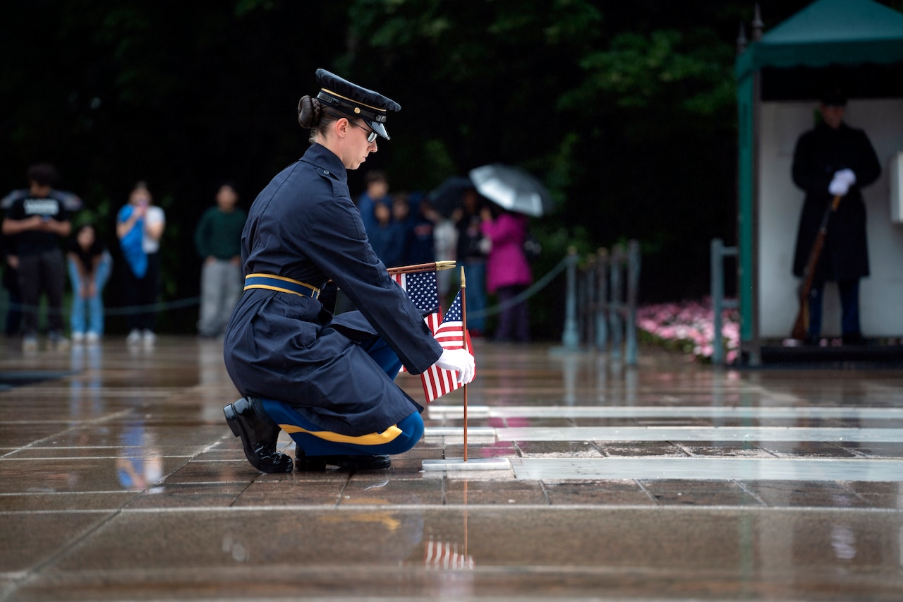 A tomb sentinel places a small U.S. flag in front of a marble gravesite during an overcast day.