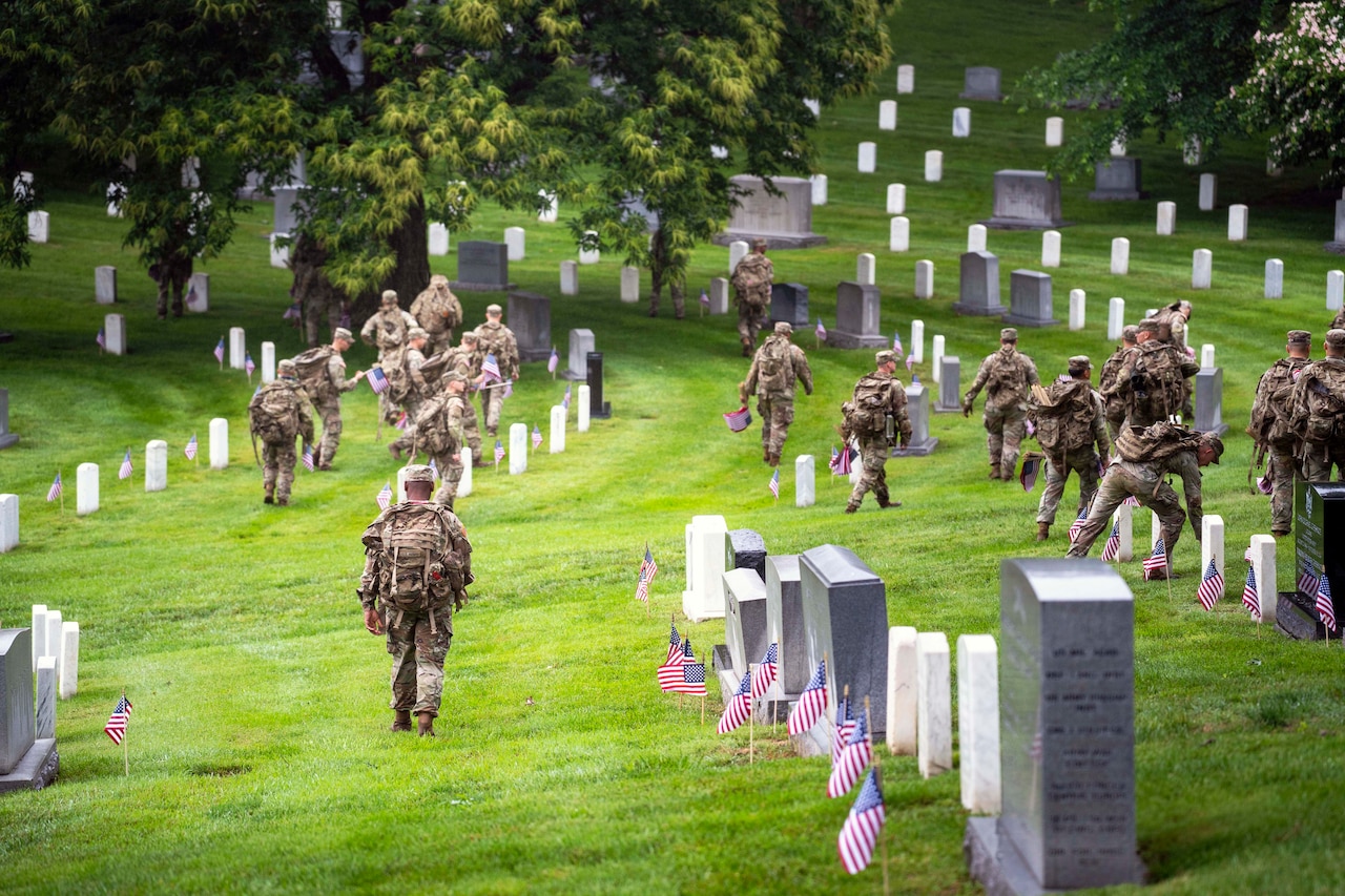 Various solders walk around a gravesite placing U.S. flags beside white tomb stones.