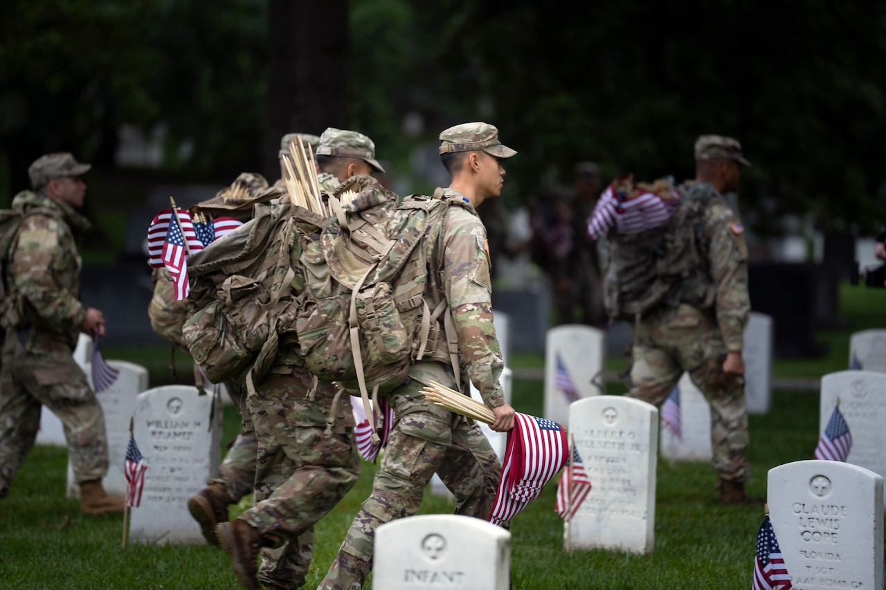 Four soldiers walk among gravestones carrying backpacks and flags during an overcast day.