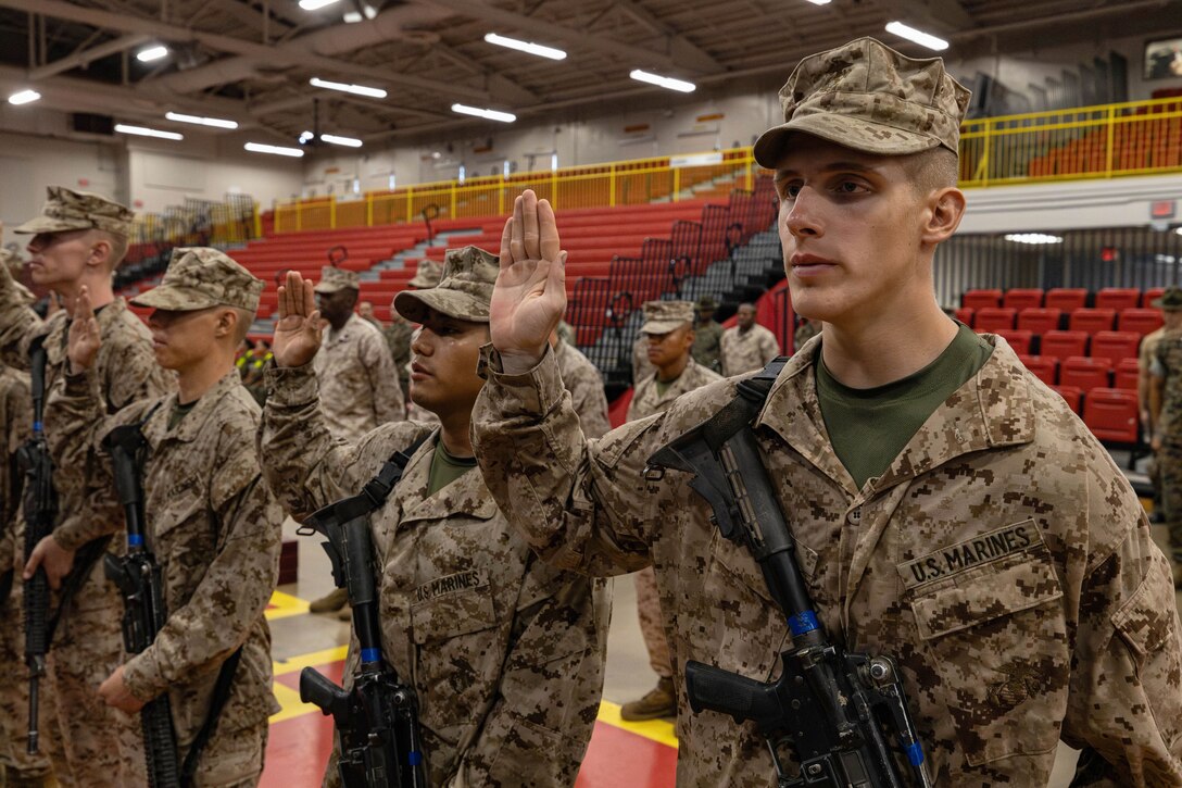Marines stand in a rows with right hands raised  in a gym-type room.