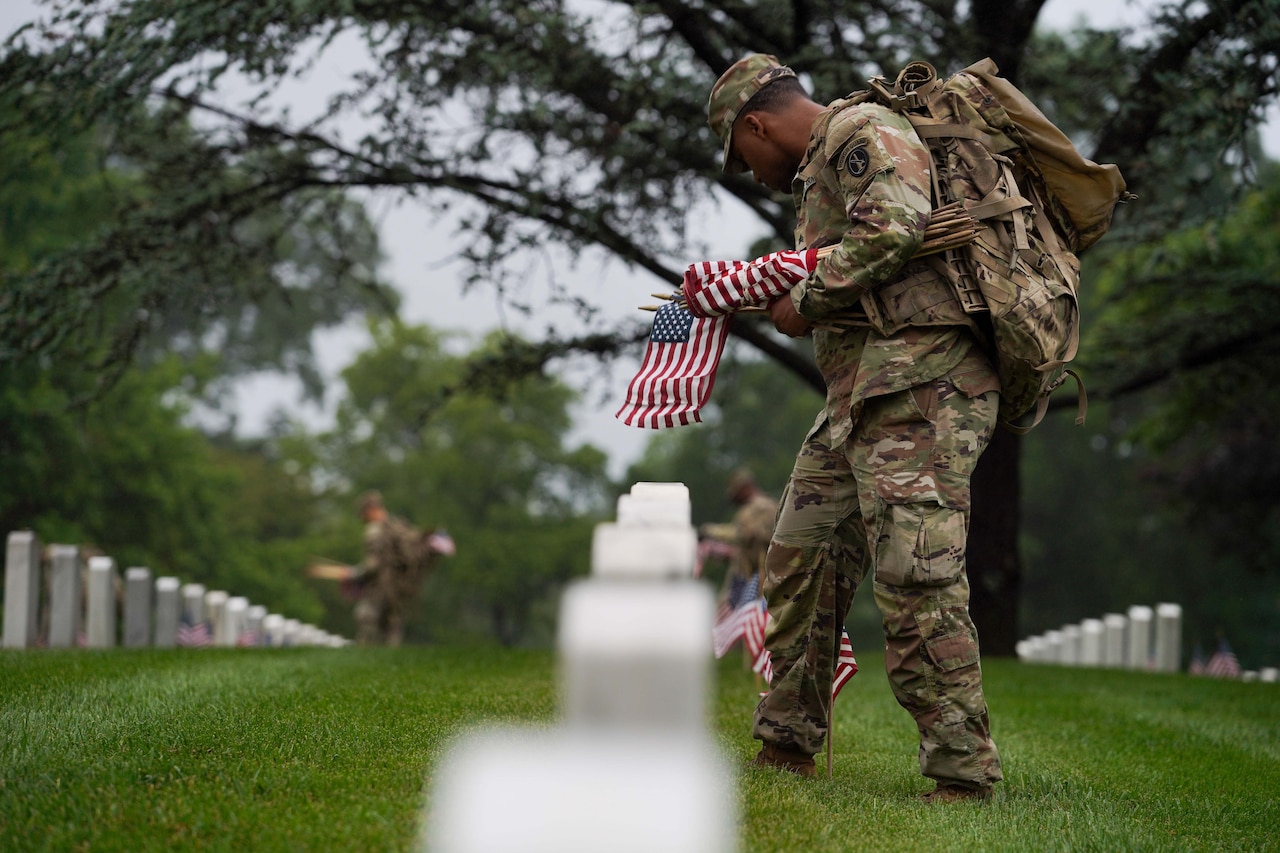 A soldier places a U.S. flag at a gravesite during an overcast day.