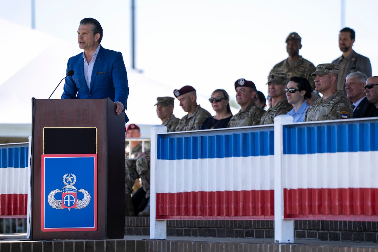 A man in civilian business attire speaks from a podium. People in camouflage uniforms and business attire are seated to the right of him.