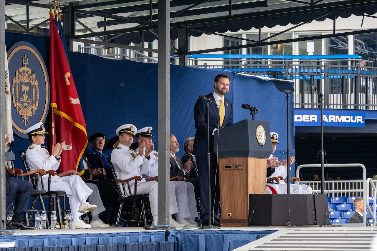 Vice President JD Vance stands at a lectern on a stage where others are seated and clap.