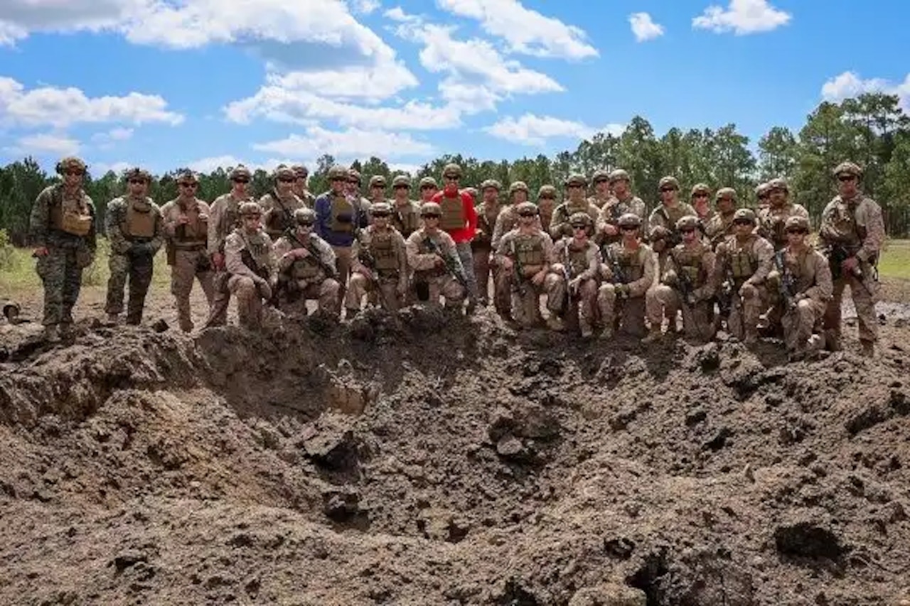 A group of Marines in camouflage uniforms and a civilian pose for a photo in front of a large dirt hole.