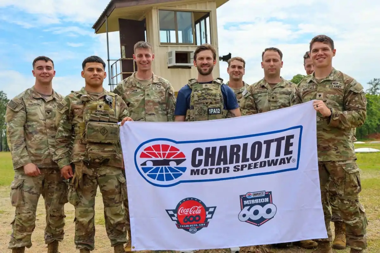 A group of soldiers in camouflage uniforms and a person in civilian clothes and camouflage vest pose for a photo while holding a sign that reads “Charlotte Motor Speedway.”