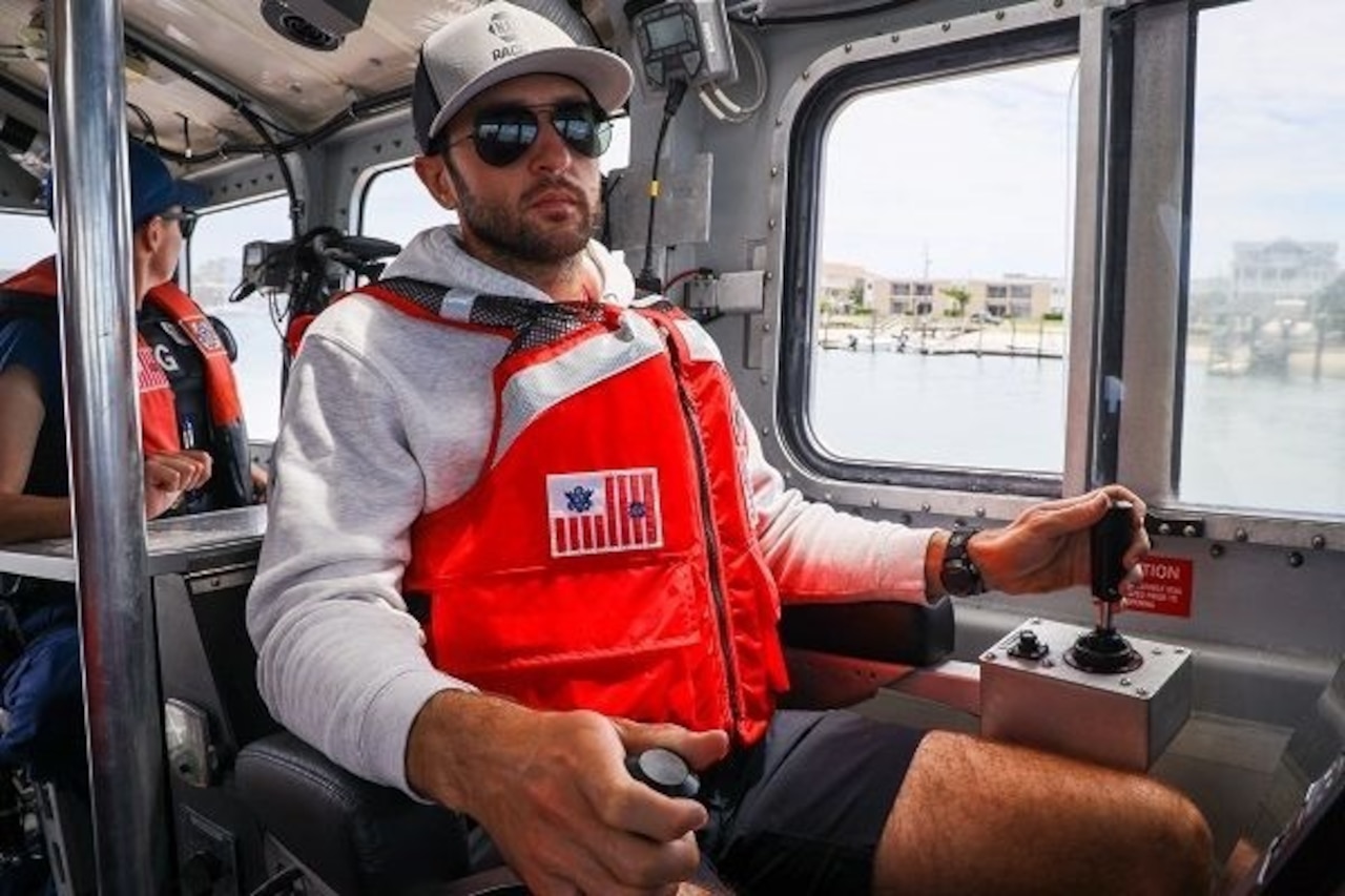 A man wearing casual attire and a life vest handles the controls of a boat. There is a person wearing a life vest looking out a window in the background.