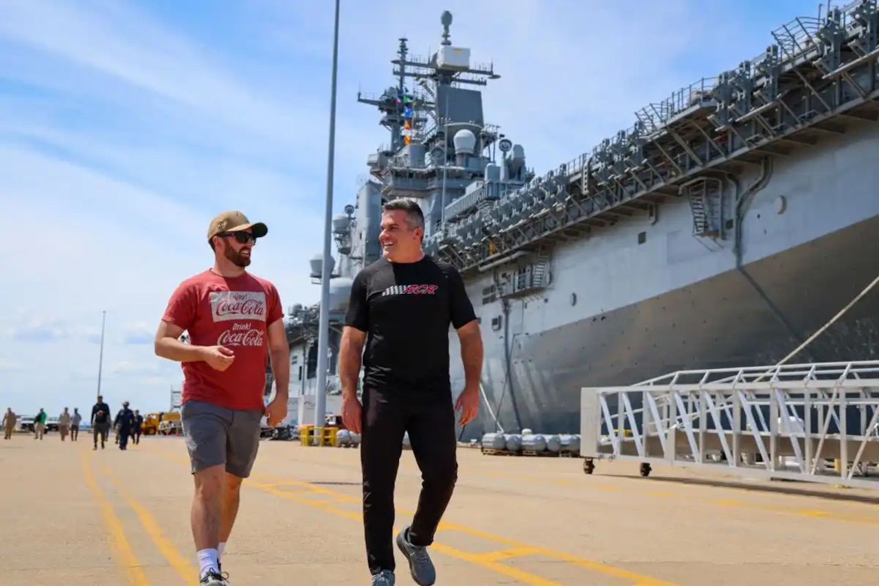 Two men in civilian attire walk down a road while a large Navy aircraft carrier is docked in the background.