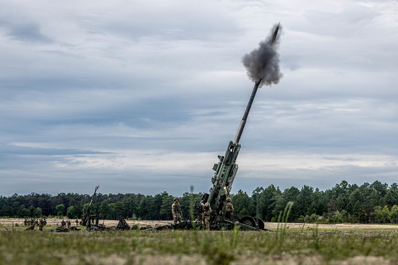 A large gun fires into the air as service members in camouflage uniforms stand around it.