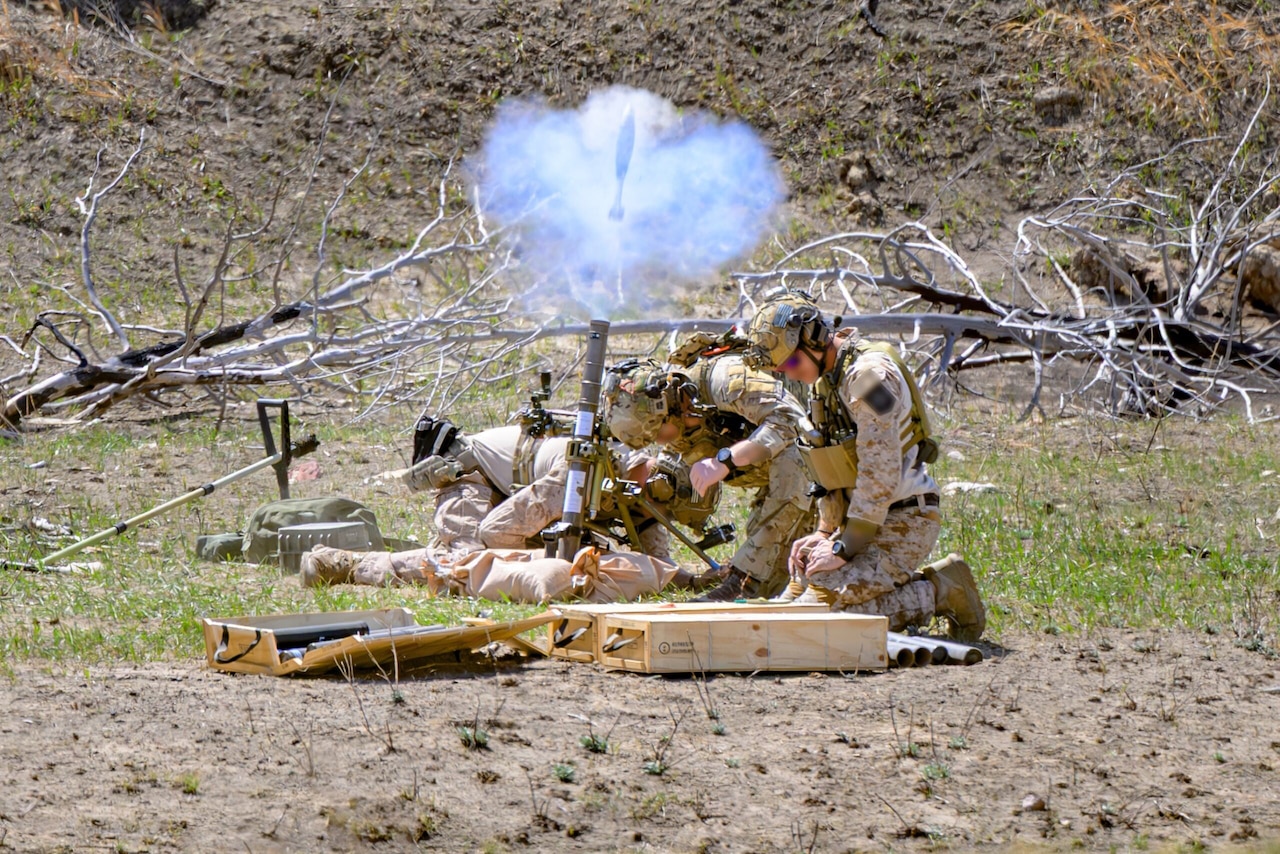 Three service members in camouflage uniforms kneel around a mortar tube and launch a round.