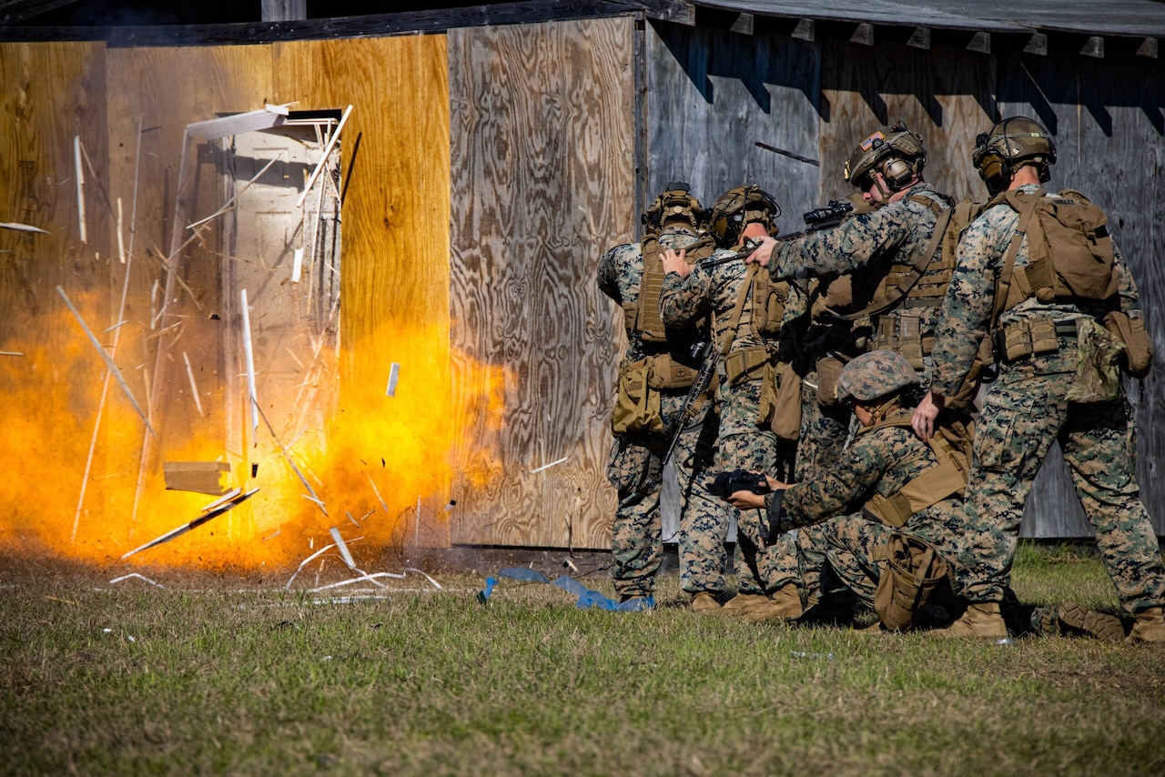 Marines in camouflage uniforms stand in formation as a door in front of them explodes.