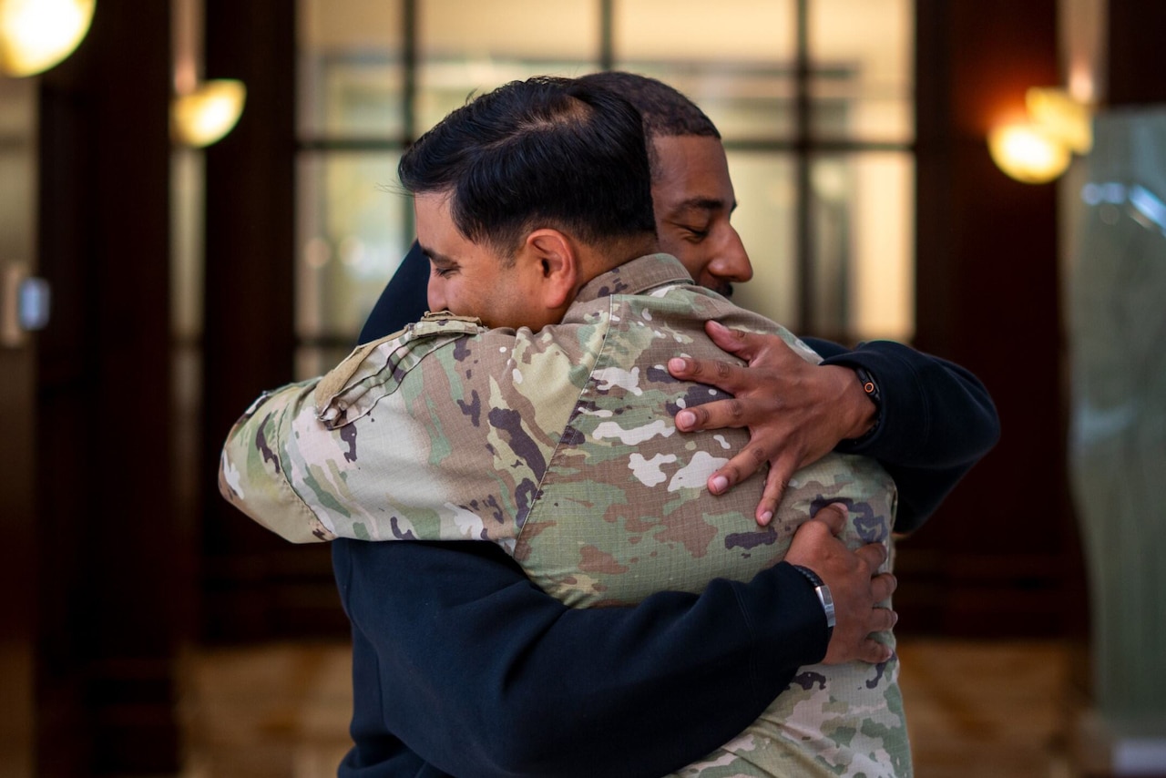 Two men in the lobby of a building hug. One is wearing casual clothes, and the other is wearing a camouflage military uniform.
