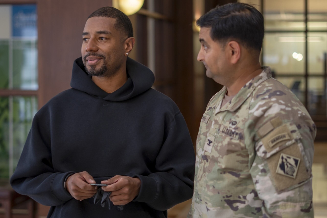 Two men are standing in a building lobby. One is wearing casual clothes and holding a flat item, and the other is wearing a camouflage military uniform.