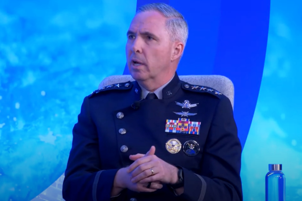 A man in a military dress uniform sits in a chair and speaks to a crowd.