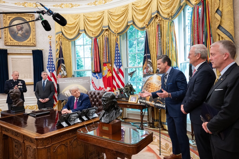 Several men in business attire stand in an oval office while one man in business attire is seated.  Another talks, with his hands outstretched.