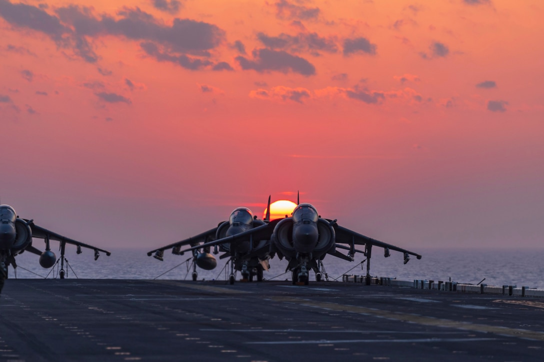 Three jets are seen on the deck of an aircraft carrier with the sun in the background in an orange and blue sky.