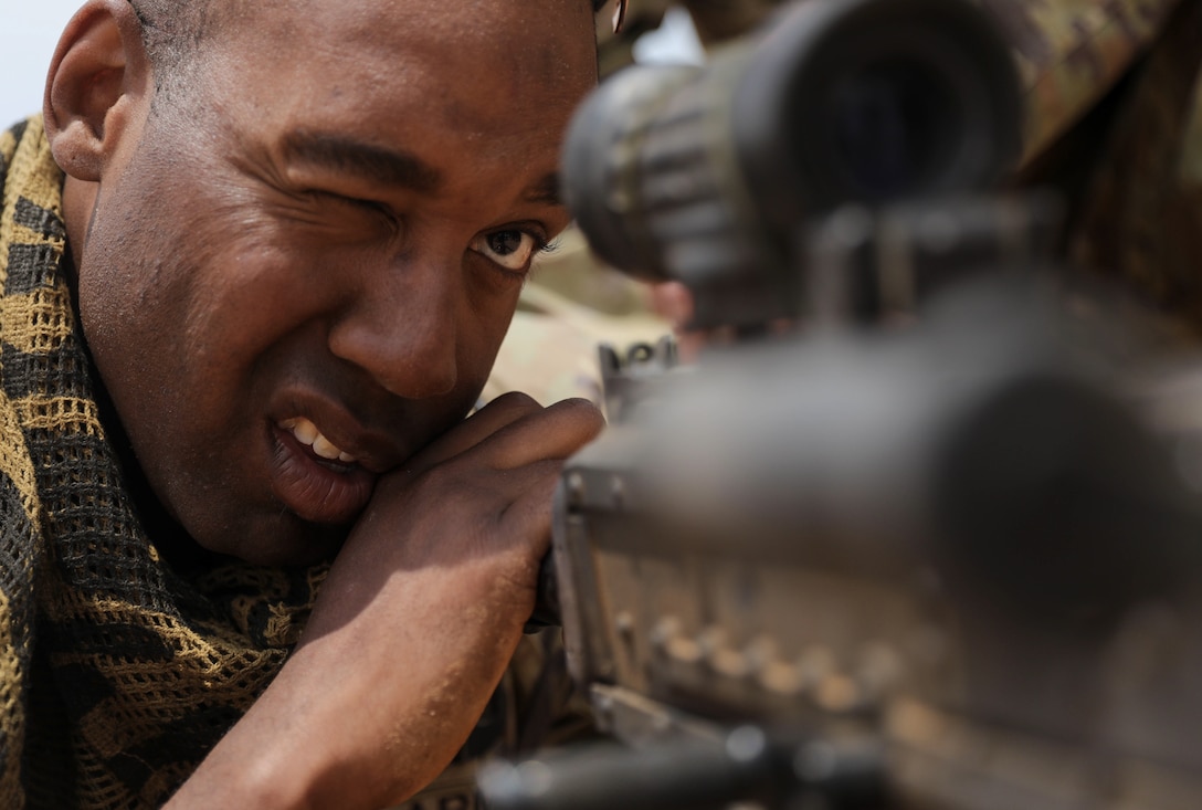 A soldier closes one eye and looks through the scope of a machine gun with an open eye outdoors.