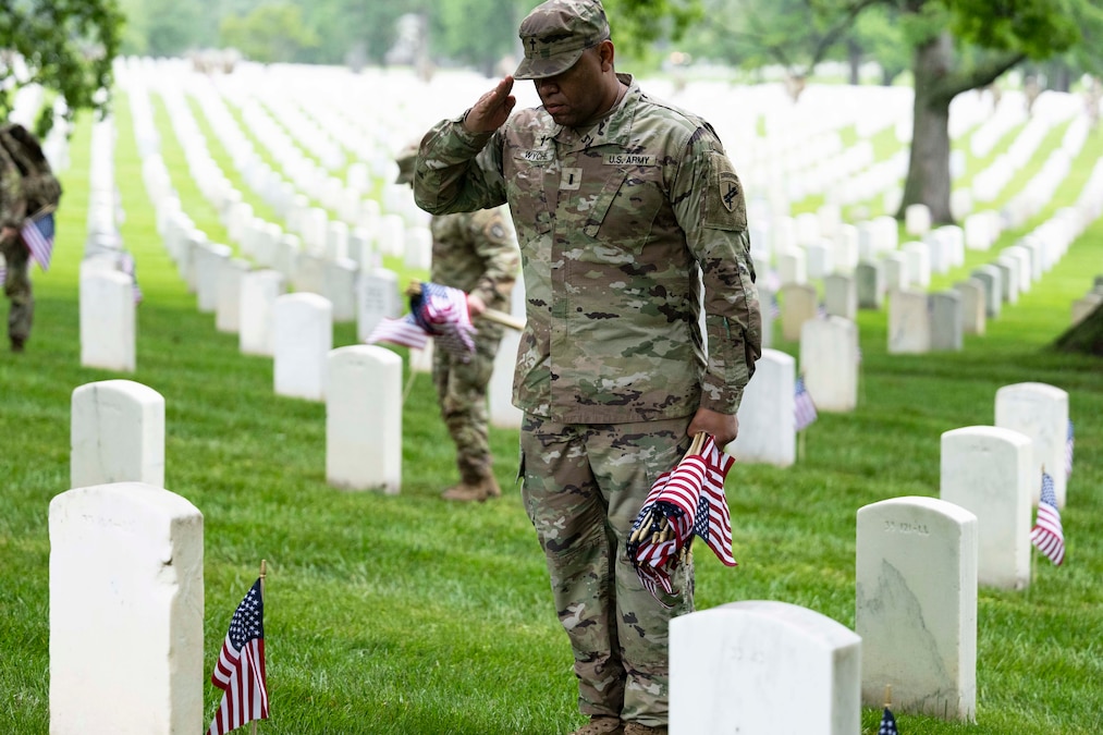 A soldier holding small American flags salutes a white gravestone with dozens of other gravestones in the background on a grassy field.